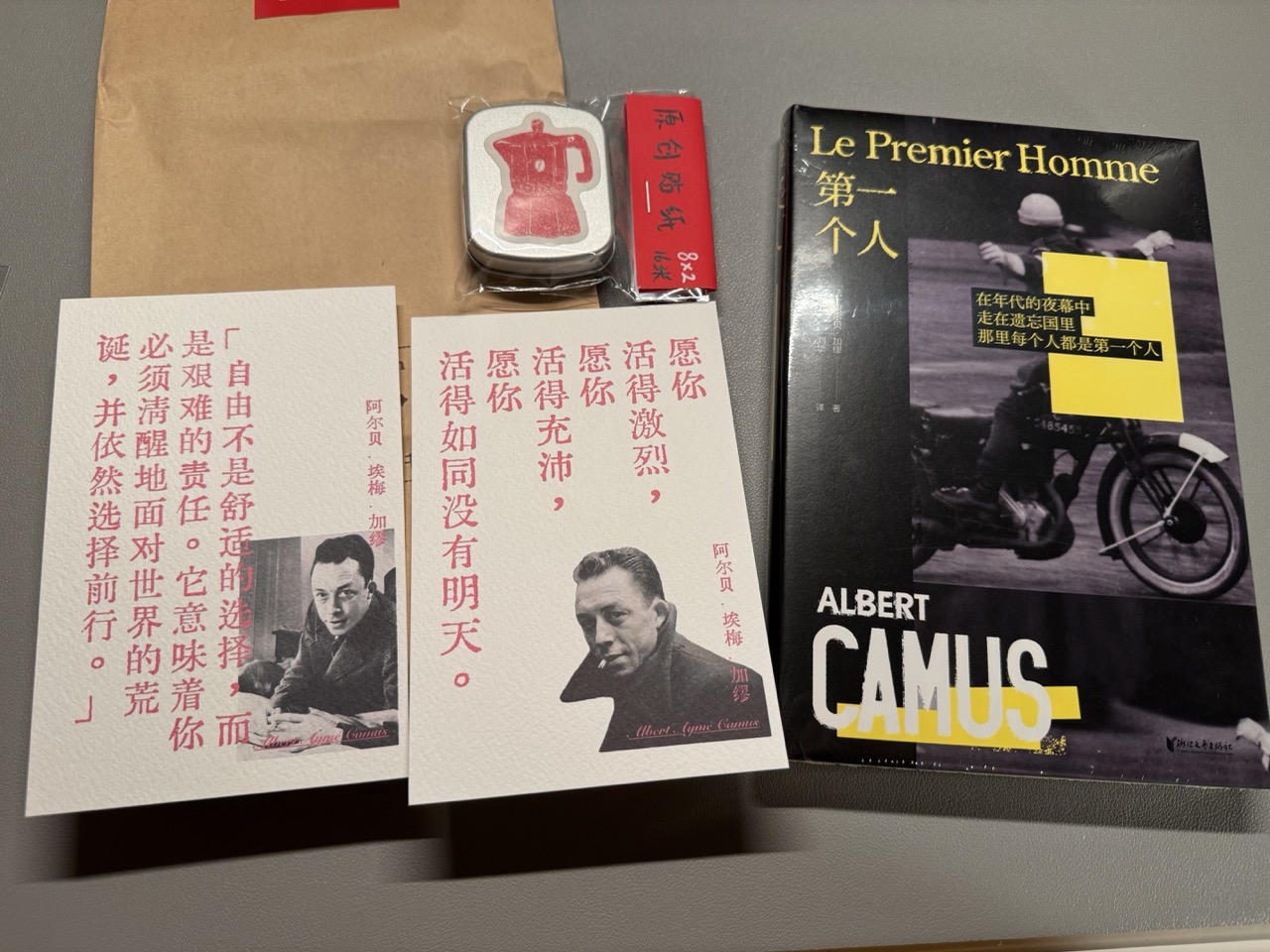 桌面上摆着加缪的《第一个人》，两张明信片上面的句子分别是「愿你活得激烈，愿你活得充沛，愿你活得如同没有明天」和「自由不是舒适的选择，而是艰难的责任。它意味着你必须清醒地面对世界的荒诞，并依然选择前行」，还有一个画着红色摩卡壶的贴纸。